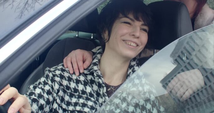 woman smiling in car seat, friend drapes arm over shoulder during playful Prague bachelorette moment, houndstooth coat, city reflections on glass, candid laughter and affectionate gesture capturing