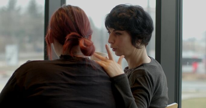 Women share private moments, Urban women converse by window, Women engage in heartfelt dialogue near cityscape, Two women exchange genuine feelings during quiet urban windowside discussion