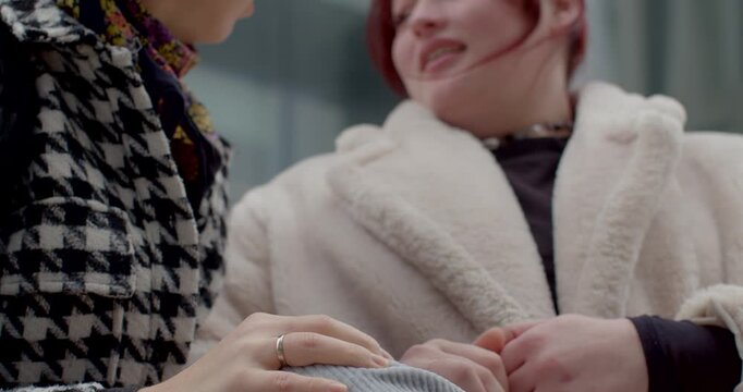 white bride-to-be and asian friend seated on bench share playful laughter and comforting hand touch, soft faux fur coat and houndstooth jacket contrast, closeup on hands and smiles, urban plaza vibe