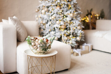 Christmas interior of a bright modern living room. New Year's 2026 trend. A bowl with snow-covered spruce branches and a baubles against a Christmas tree.