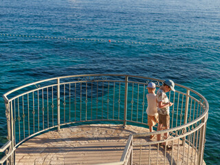 Two boys standing on seaside terrace with iron railing looking at calm blue water, copy space. Concept of childhood exploration, family travel, summer holidays.