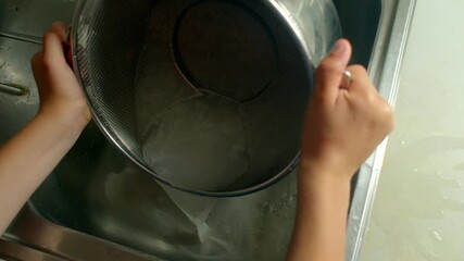white woman washing large metal pot and colander in sink, running water from faucet, scrubbing and rinsing, domestic