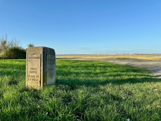 Vintage stone direction post on the Somme Bay foreshore pointing to Cap Hornu, the Upper Town and Chapelle Saint-Valery, set amid green saltmarsh with distant Le Crotoy under a clear blue evening sky