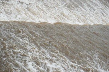 The flow of river water is very fast from a dam, the water is brown and foamy, indicating a strong...