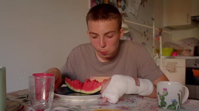teen enjoying watermelon slice at patterned tablecloth, juicy summer snack, bandaged hand resting nearby, glass and mug