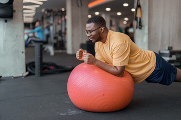 Bright indoor gym scene with man exercising on red stability ball smiling focused