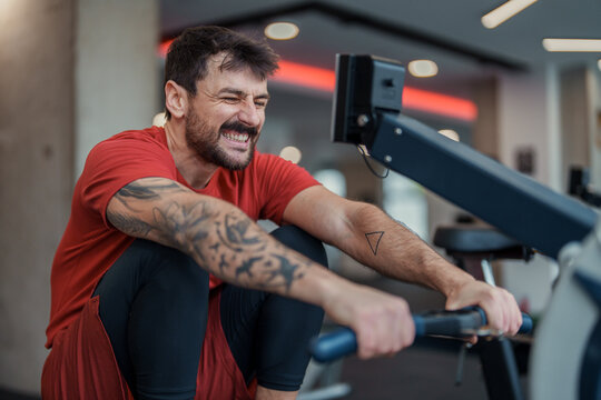 Focused Athletic Man in Red Shirt and Black Leggings Exercising on Rowing Machine in Gym - Powered by Adobe