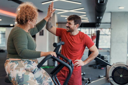 Middle aged woman exercises on stationary bike with young male trainer in bright modern gym setting