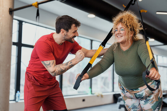 Professional Personal Trainer In Red Shirt Guiding Young Woman Through TRX Suspension Training Workout In Contemporary Fitness Center
