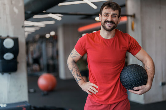 Smiling Tattooed Male Athlete Poses With Exercise Ball In Modern Fitness Center