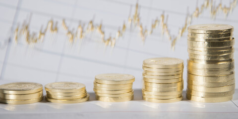 Neatly stacked gold coins arranged in ascending height with a financial graph blurred in the background.