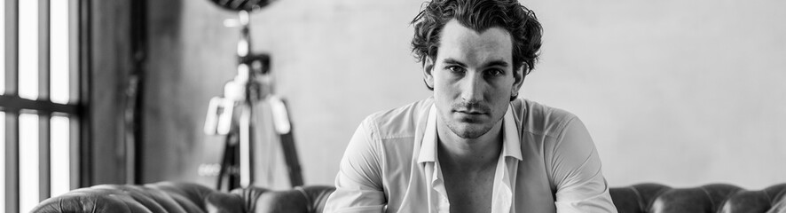 Banner. Young sexy handsome fit man in a brown suit looking at the camera sitting on a chair in the studio. Grey background, loft interior, studio. Causal stylish men's clothes, watch