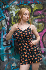 Blonde sensual woman with long legs posing confidently in front of a colorful graffiti wall, wearing a short floral dress. Urban fashion meets vibrant street art in this expressive portrait.