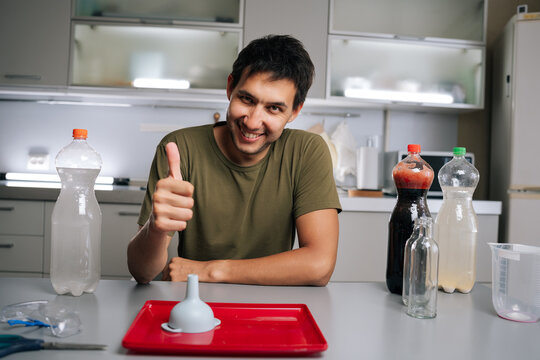 Cheerful gardener giving thumbs-up gesture to indicate success or approval sitting at table in kitchen, preparing fertilizer for plants using various liquids and funnel, smiling looking at camera.