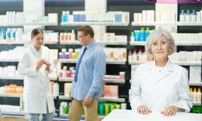 Obraz premium Positive old female pharmacist posing in chemist's shop with large assortment of goods