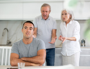 Fototapeta premium Family of threen - mother, father and son quarrel in kitchen. Eldery cuple is disappointent of adult son.