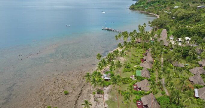 Savusavu resort with traditional bures huts and lush tropical landscape reef coastline. Exotic relaxing travel vacations and scenic views of the tranquil bay with boats and a long pier. Aerial flight