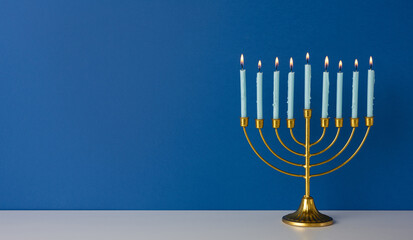 Obraz premium A golden menorah with nine lit candles sits on a white surface before a deep blue wall, evoking Hanukkah tradition, warmth, and holiday celebration