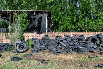 A large pile of discarded and polluting tires in front of an abandoned warehouse with a damaged roof and overgrown plants. Industrial site, waste, pollution, environment, recycling, ecology, eco