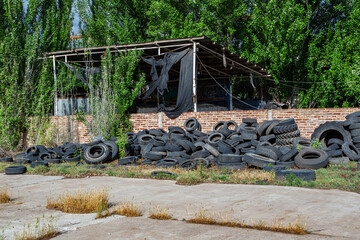 A large pile of discarded and polluting tires in front of an abandoned warehouse with a damaged roof and overgrown plants. Industrial site, waste, pollution, environment, recycling © robscri