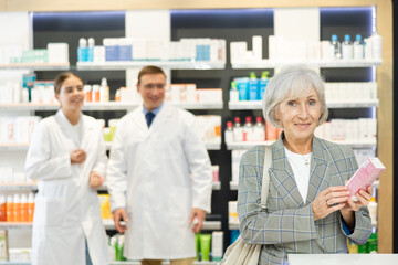 Obraz premium Near self-service window in pharmacy, senior female customer chooses cream for aging skin, carefully reads information for consumer on packaging of product