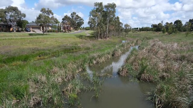 A calm stretch of Skeleton Creek bordered by reeds and grasslands in Melbourne&rsquo;s western suburbs. Concept of urban wetland ecology, water management, and coexistence of residential living