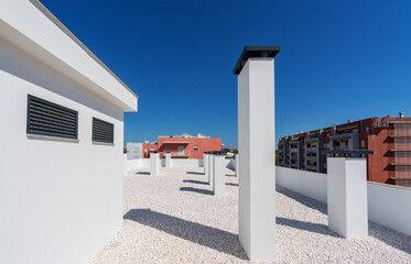 A rooftop view featuring several white chimneys against a vibrant blue sky, surrounded by modern buildings in a sunny urban setting © Sergo