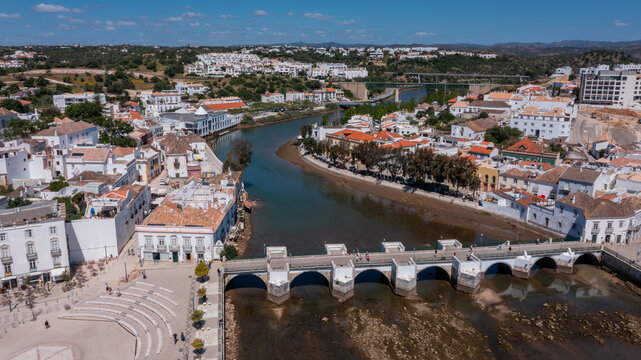 Aerial view of Tavira with historic Roman bridge over the Gilao River, traditional buildings and green hills in background under blue sky in Algarve, Portugal. Concept of cultural heritage tourism