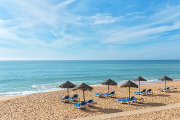 Sun-kissed sandy beach features lounge chairs arranged beneath thatched umbrellas, inviting relaxation by the calm ocean waves and clear blue skies