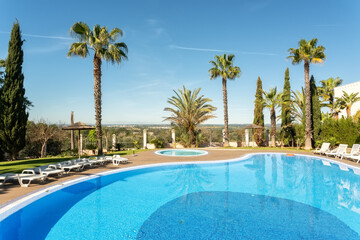 A serene scene shows a large swimming pool surrounded by palm trees at a villa on a sunny afternoon with clear blue skies. Guests can relax and unwind by the water