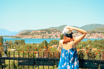A woman in a flowing blue dress and sun hat stands on a balcony, overlooking the stunning Lake...
