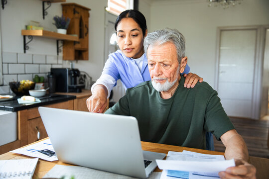 Adult caregiver helping senior man with online bills at home, focused