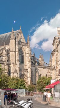 Basilique Saint-Michel timelapse in Bordeaux, France, is a gothic landmark with a tall spire. People enjoy cafes nearby, under green trees and blue cloudy skies, capturing the city's vibrant charm