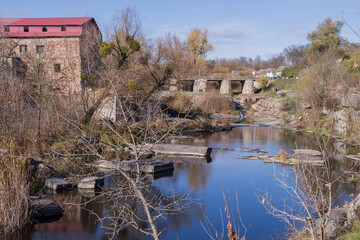 Scenic autumn view of a historic stone mill, old bridge and rocky river with reflections, dry vegetation and rural nature. Peaceful landscape with heritage architecture.