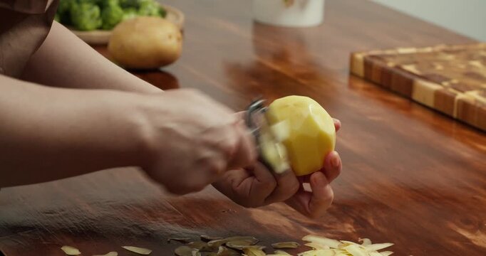 Close up of hands peeling fresh potato with peeler on wooden table, preparing ingredients for homemade vegan meal, cooking process, healthy food, rustic kitchen atmosphere, natural lifestyle
