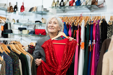 Elderly woman buyer chooses evening dress in clothing store..
