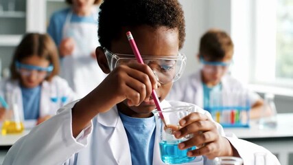 A young boy in a white lab coat and goggles holds a pencil and beaker filled with blue liquid in a science class setting, surrounded by equipment. He is focused on the task. - Powered by Adobe