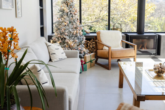 Frosted Christmas tree standing near windows and glowing with warm white lights above wrapped gifts