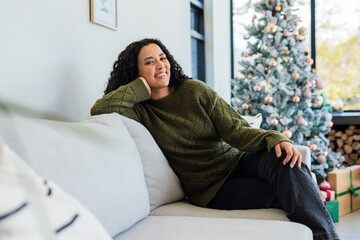 African American woman reclining on gray sofa at home smiling at camera near frosted Christmas tree