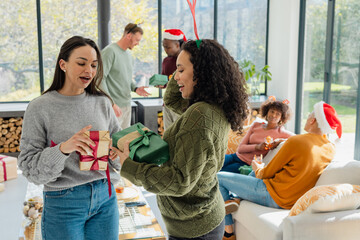 Diverse female coworkers wearing headband exchanging wrapped gifts in living room with glass table