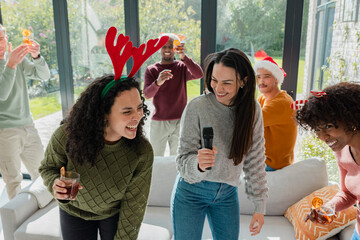 Diverse friends singing on sofa holding cocktails and mic wearing festive headwear in living room
