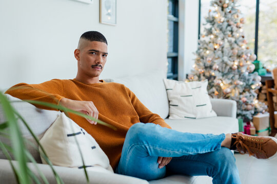 African American man sitting on sofa at home wearing orange sweater beside Christmas tree