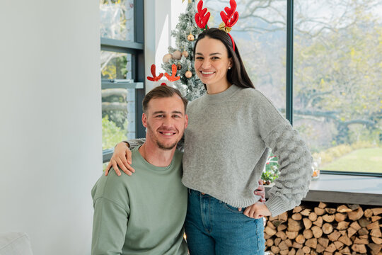 Couple posing and smiling by Christmas tree and firewood in living room wearing antler headbands
