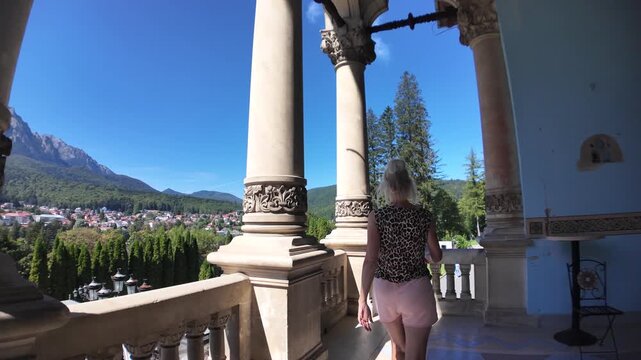 Woman tourist on Cantacuzino Castle balcony in Busteni, Romania, gazing over mountain peaks and the historic courtyard under a clear blue summer sky, enjoying travel discovery in Transylvania