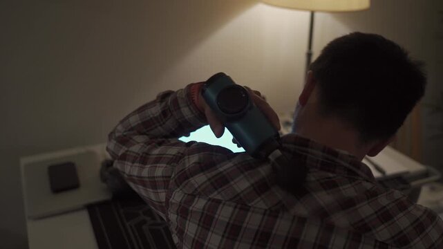 Neck pain, cervical osteochondrosis. Male student studying until night at computer with neck pain. Tired guy kneads neck with pistol shock massager while working on laptop. Muscle recovery and massage