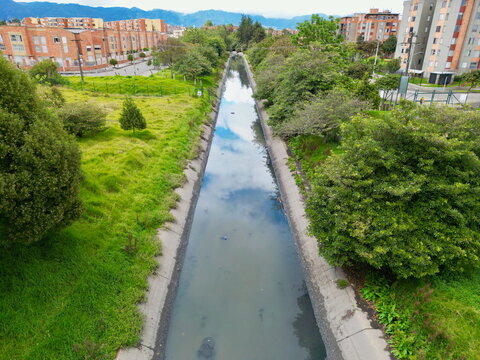 Low aerial drone view of the straightened, polluted canal of the Quebrada La Salitrosa flowing through a dense urban residential area near Suba, Bogot&aacute;, Colombia.