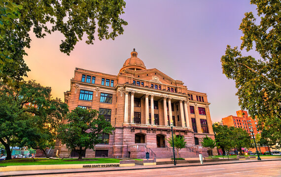 Historic Harris County Courthouse of 1910 in Houston, Texas, at sunrise. The granite building and dome are lit by warm light under a colorful sky, framed by trees