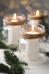 Christmas lanterns. Burning candles in glass jars and fir tree branches on white table against blurred lights, closeup