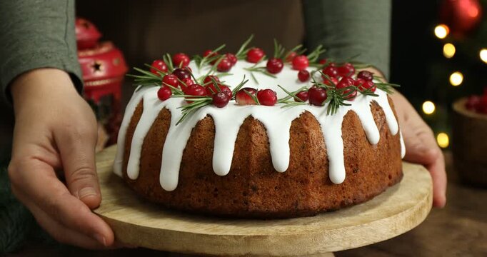 Woman putting delicious Christmas cake with icing, cranberries and rosemary on wooden table against black background with blurred lights, closeup