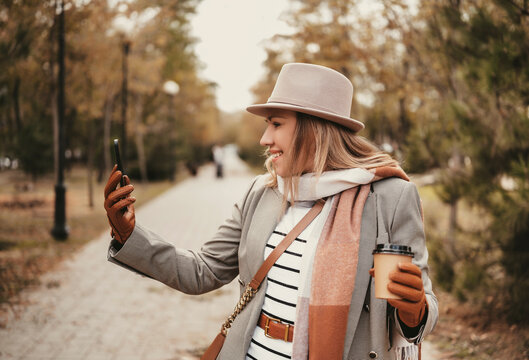 Blonde woman in business attire enjoying autumn day outdoors using her smartphone and sipping coffee - Powered by Adobe